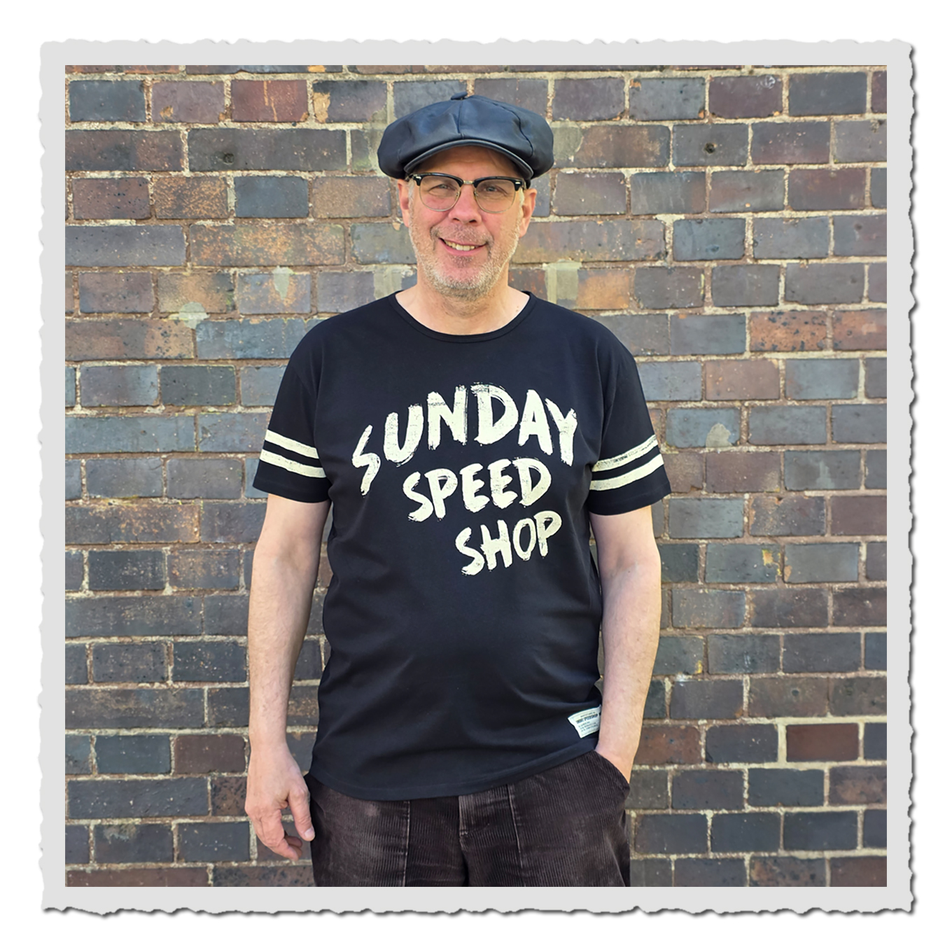 Person von vorn mit schwarzem Sunday SpeedShop Shirt vor gemauerter Backsteinwand.